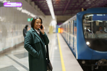 Obraz premium Happy successful businesswoman returning home from office talk on smartphone wait for train at subway station. Smiling african woman call to family or husband from underground after long working day