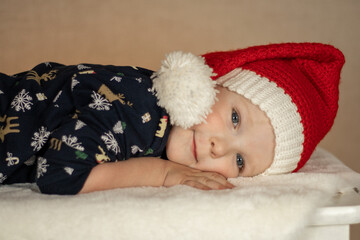 a little cute boy in a santa hat and New Year's pajamas lies on a fur rug on a plain background. close-up