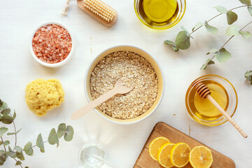 Natural ingredients for homemade scrub. Oat flakes, oil, honey, pink himalayan salt, lemon, face or body brush, sponge and eucalyptus branches on white wooden table. Top view, flat lay