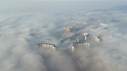 Silhouettes of tall houses through the incoming thick fog. Megalopolis in white clouds in the early morning at time.