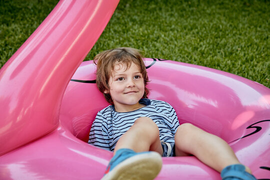 Blond boy in blue clothes, lying on pink inflatable flamingo on grass.