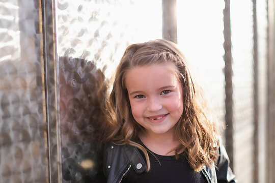 Smiling young girl with dimples leaning against a metal wall.
