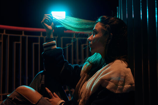 A pensive young woman is illuminated by a blue light at night