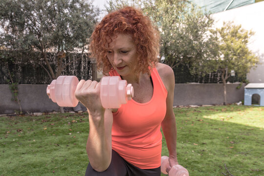 Mature redhead woman lifting dumbbells at garden, home fitness