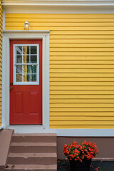 Fototapeta premium The exterior facade wall of a yellow wood clapboard siding house. The door of the building is bright red with a small glass window. Steps lead up to the door with a pot of red flowers on the ground.