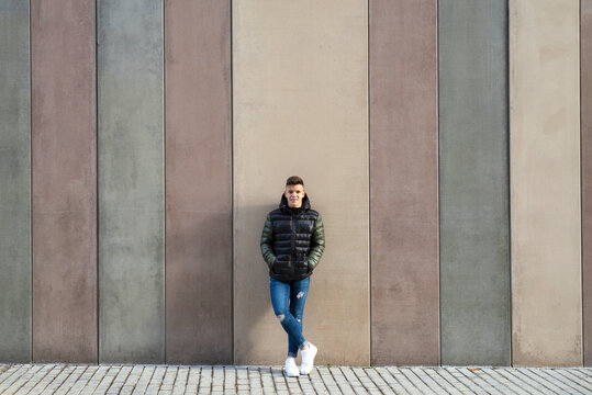 Happy young guy leaning on colored wall hands on pocket,looking camera