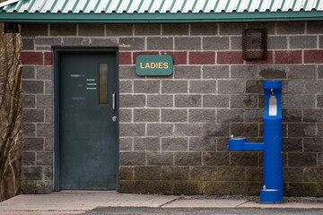 The entrance to a ladies washroom in a park. The lavatory has a green door, brick wall, wooden...