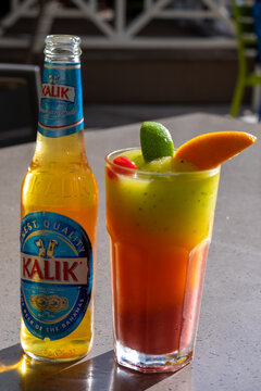 Orlando, Florida, US- December 2021: A tall clear bottle of Kalik beer from the Commonwealth Brewery in the Bahamas. The pale lager ale on a patio table next to a tall fruit punch with fruit wedges.