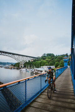 A young man bikes across a bridge with water and boats in the distance