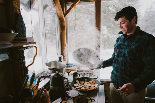 A young man cooks steaming breakfast in a cabin during winter
