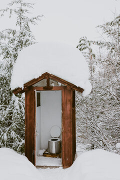 An outhouse with toilet seat up covered in deep snow in the woods