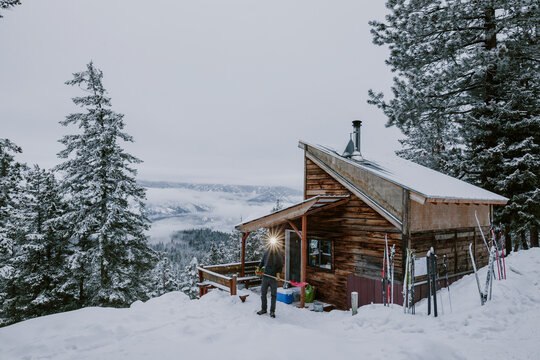 A man with headlamp shovels snow in front of cabin and mountain view