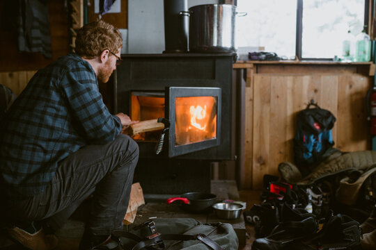 A man wearing flannel stokes the fire inside a ski cabin during winter