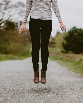 A woman with leather boots jumps in the air on a gravel trail