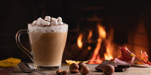Cocoa with marshmallows and chocolate in a glass mug on a wooden table near a burning fireplace, horizontal banner