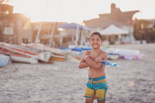Boy at the beach with arms crossed holding water goggles.