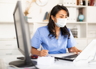 Hispanic woman general practitioner wearing medical mask working with case histories on laptop in modern office.