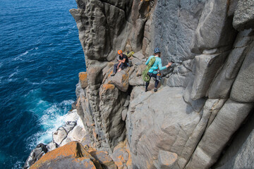 A couple of intrepid rockclimbers explore the featured sea cliffs of Cape Raoul, smiling at each other as they traverse the exposed terrain above a seal colony, in Tasmania, Australia.