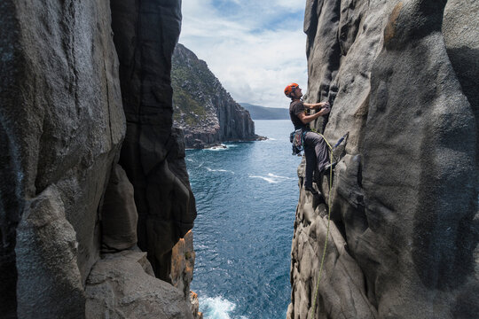 Daring man rockclimbs the exposed edge of a dolerite rock ridge, with the ocean seen in the background in Cape Raoul, Tasmania, Australia.