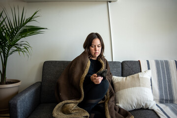 Woman wrapped in blanket sitting on couch looking at phone