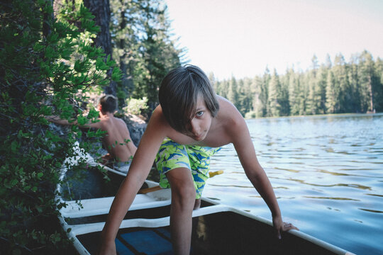Boy in Shark Shorts Steps Into Canoe on a Lake Edge. Summer Day.