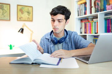 Young man studying at home with laptop, papers and book on desk. Distance learning concept.