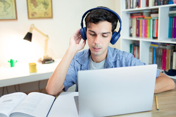 Young man studying and listening on headphones  at home with laptop, papers and book on desk. Distance learning concept.