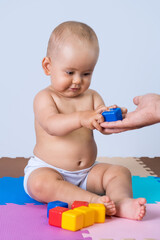 The toddler takes a blue plastic cube from his father's hands. A newborn baby in the playroom, sitting on a soft puzzle floor