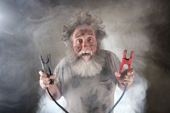 Crazy old man with beard holding jumper cables surrounded by smoke receiving a shock