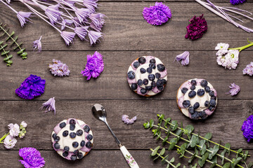Three blueberry Graham Cheesecakes topped with fresh berries with a spoon on a dark wood background with purple flowers and twigs. Flat lay.