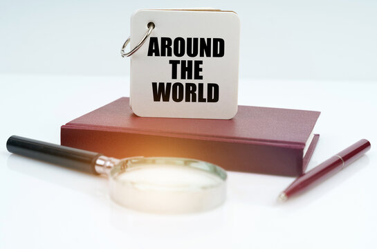 On a white surface lies a red notebook, a pen, a magnifying glass and a notebook with the inscription - Around the World