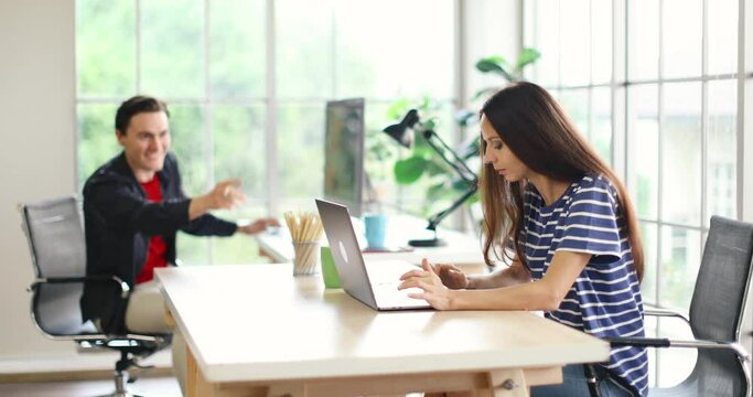 Millennial Caucasian young female worker in casual outfit sitting typing working with laptop computer while happy funny male colleague freelancer inviting her listening to music and dance together