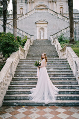 Fototapeta na wymiar Bride in a long dress stands half-wrapped on stone steps near an old building