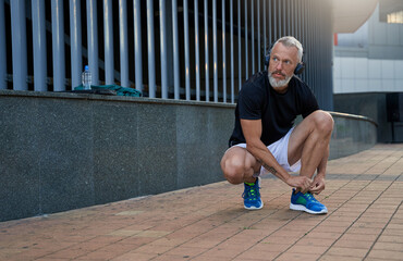Middle aged sportsman looking away while tying shoelaces on his blue running sneakers, getting...