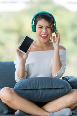 Close up shot portrait of young beautiful Asian woman sitting on sofa  and listening to music with headphone in relaxing manner and hold smarthphone in hand. Green nature blur in background