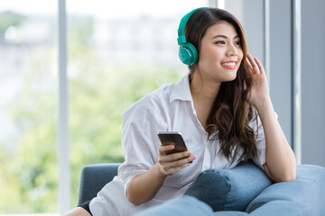 Close up shot portrait of young beautiful Asian woman sitting on sofa  and listening to music with headphone in relaxing manner and hold smarthphone in hand. Green nature blur in background