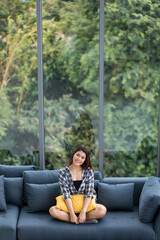 Asian woman sitting on sofa near big glass windows, relaxing alone in house with green forest in background.