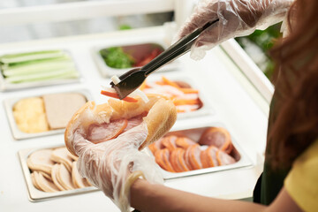 Closeup image of cook using tongs to put fresh vegetablesn sandwich of customer