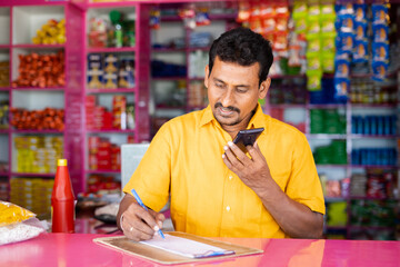 Indian groceries business man noting orders while talking with customer on mobile phone at kirana...