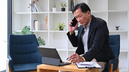 Naklejka premium Mature businessman talking on mobile phone and working with computer tablet in his office room.