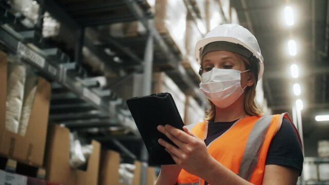 Warehouse worker wearing medical mask, safety vest and hardhat standing near shelves with goods using digital tablet checking inventory in stock room of a manufacturing company. E-Commerce concept.