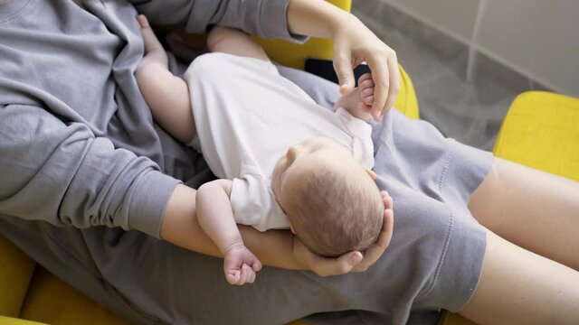 Mother with sleeping baby relaxing at home. Detail view unrecognizable woman cradles cute child on knees while sitting in large cozy armchair in modern apartment, tracking shot