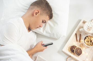Obraz premium A teenage boy in a white T-shirt lies in bed with a smartphone in his hands. Next to it is a tray of Christmas breakfast. Digital addiction. A cozy festive morning. Bed vibes