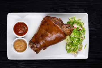 Tasty pork knuckle with sauces and vegetables on a black wooden background