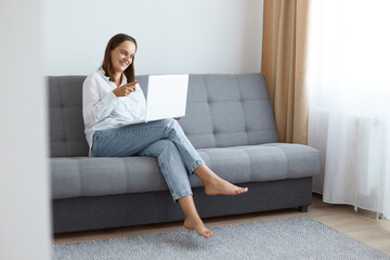 Indoor shot of smiling happy young adult woman wearing white shirt and jeans sitting on sofa at home and working on laptop, having online conversation, pointing finger to device camera.