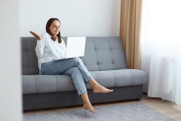Confused young adult woman wearing white shirt and jeans sitting on sofa at home and working on laptop, having puzzled facial expression, showing helpless gesture.
