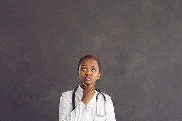 Pensive African American female healthcare worker holding her chin and looking up thinking about diagnosis. Serious woman in medical gown on gray concrete background. Medicine concept. Place for text.