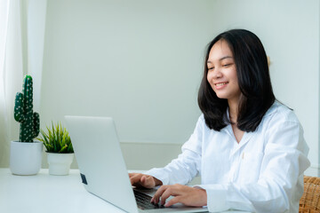 Portrait Cute Asian teen with long hair Wearing a white shirt, sitting happily studying online with a laptop on the bedroom desk.