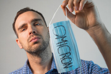 A sad European man with a small beard wearing a blue plaid shirt is holding a medical mask with...