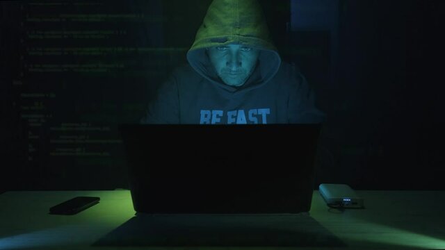 Male Hacker In Hood Working On Laptop In A Dark Room At Night. - zoom in